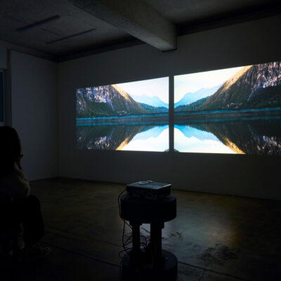 Photograph of two video stills projected onto a wall in a dark room. The images are mirrored and show a body of water with mountains reflected onto it in an almost butterfly shape