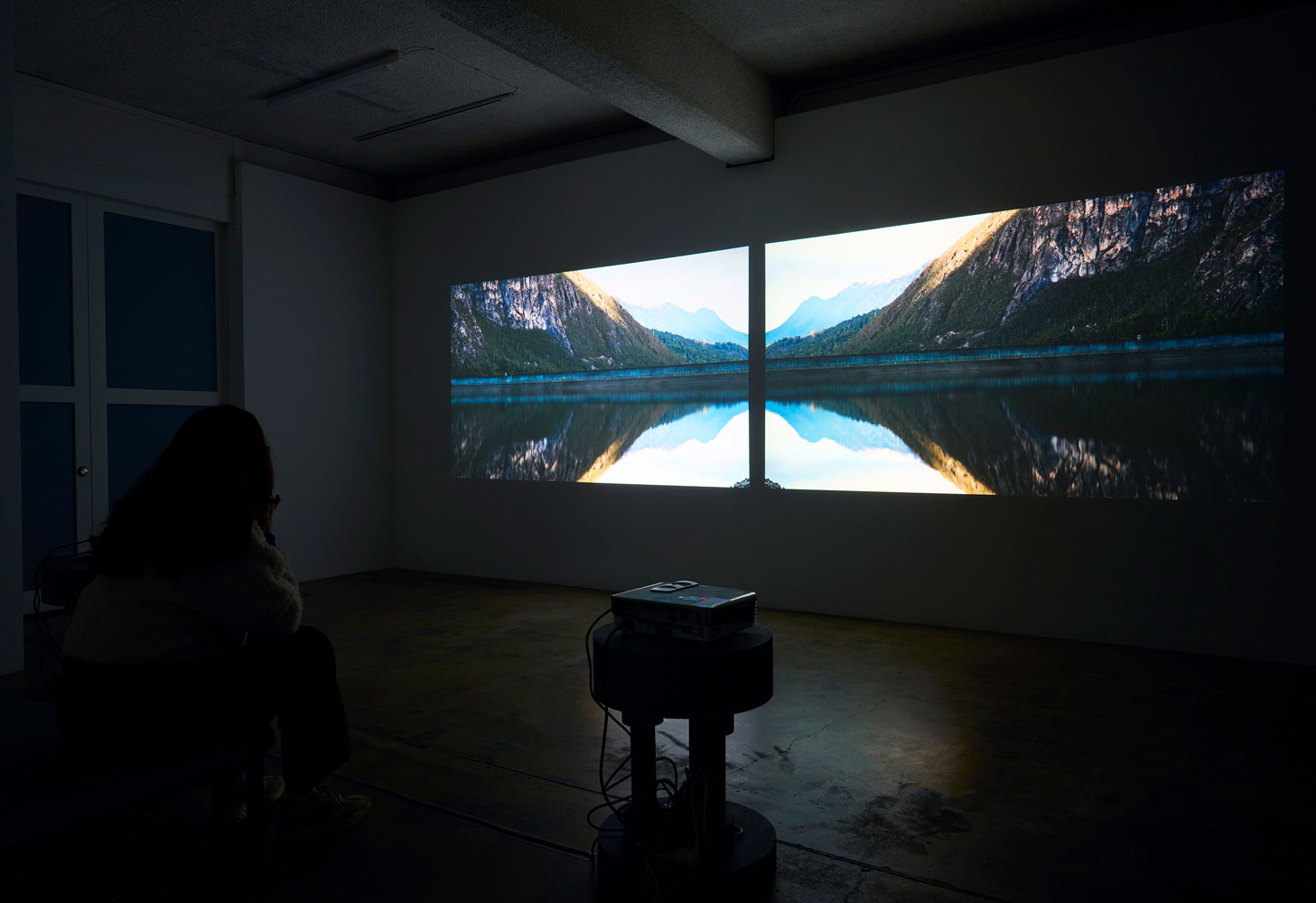 Photograph of two video stills projected onto a wall in a dark room. The images are mirrored and show a body of water with mountains reflected onto it in an almost butterfly shape
