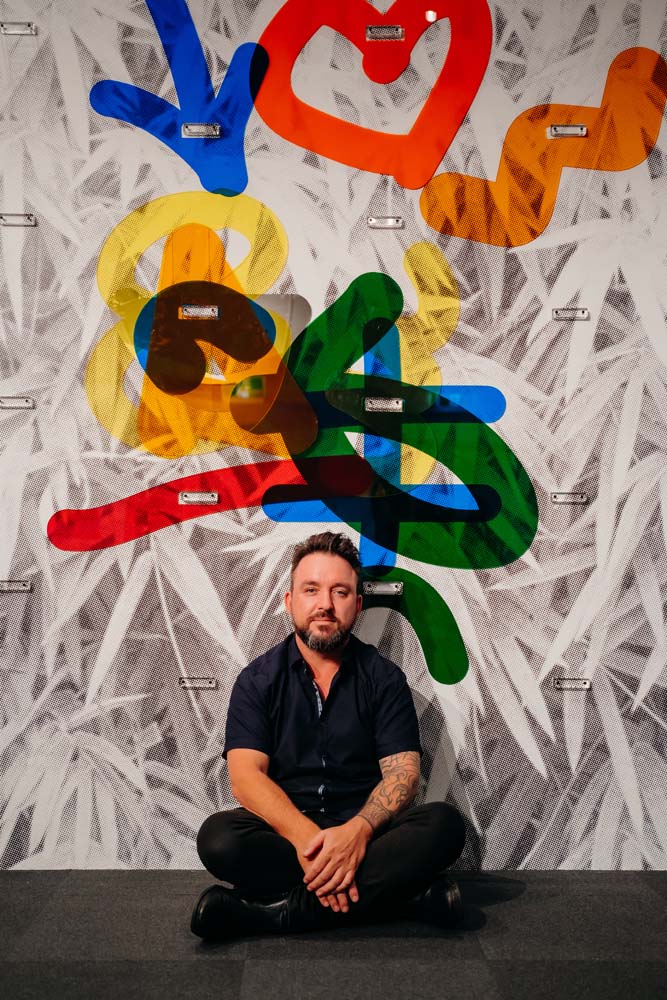 A man sitting in front of an interactive artwork. It is a black and white wall vinyl background with colourful acrylic cut out pieces that can be moved and placed into other areas. The man is sitting cross-legged on the floor.